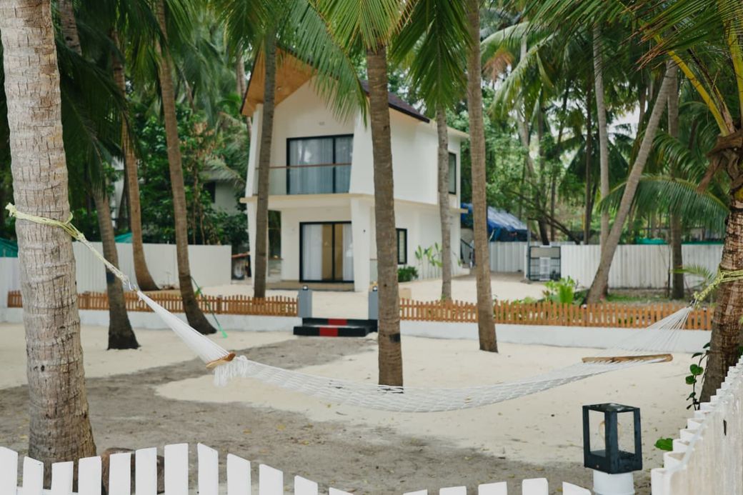 A white two-storey beach cottage among coconut palms, with a hammock slung between the palms on the sand
