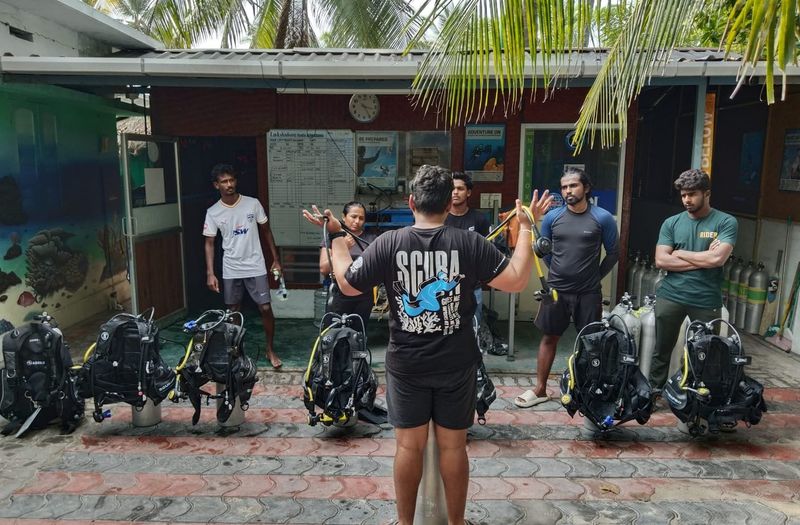 An instructor briefing six divers in the yard outside the Lak Scuba dive centre, eight assembled scuba units lined up on the ground between them