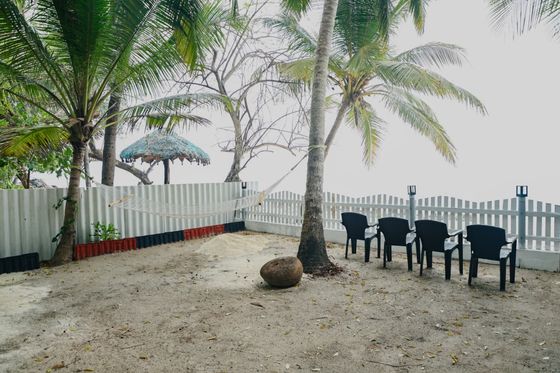 Four black chairs and a hammock on the sand inside a white fence, under coconut palms, with a thatched sunshade beyond