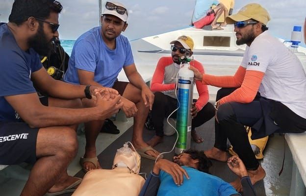 An emergency scenario on the dive boat: instructors and students around a casualty and a CPR manikin, with an emergency oxygen cylinder and mask in use