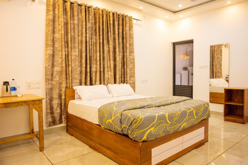 A Standard AC room: a double bed with a grey and yellow quilt, a small wooden table with a flask and a water bottle, and a mirror beside a glass door