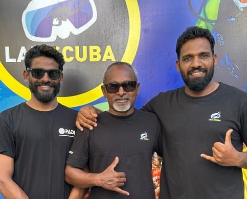 Three of the Lak Scuba team, in branded PADI and Lak Scuba T-shirts, standing in front of the dive centre's painted LAK SCUBA sign