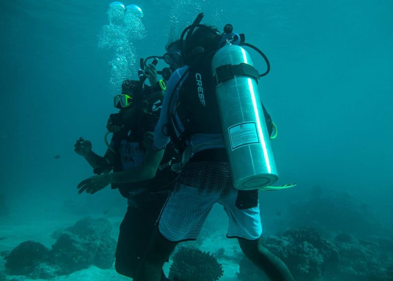 Two divers descending together in blue water over the reef, one close in front of the other, their bubbles rising