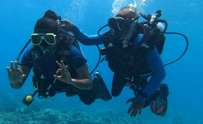 Two scuba divers underwater making the "OK" hand sign, wearing full scuba gear and surrounded by clear blue water.
